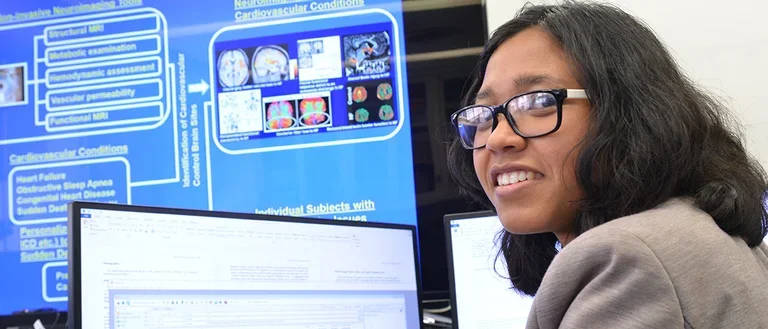 A Kumar Lab member is sitting at a computer screen giving a presentation. A larger blue screen with colorful brain scans is in the background.